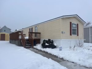 A beige mobile home with red shutters and a wooden porch is shown on a snowy day, with snow covering the ground and steps.