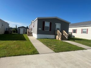A gray manufactured home with white trim and red shutters, two wooden staircases, and a small shed in the backyard, set on a grassy lot with a concrete walkway.