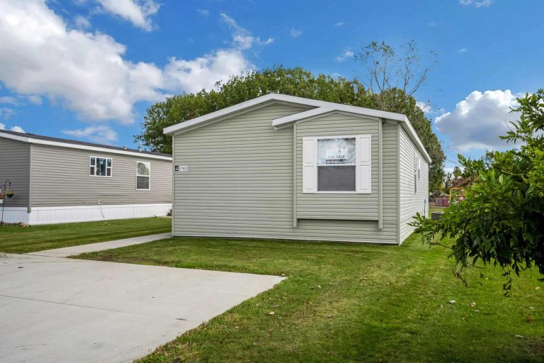 A light gray manufactured home with white trim sits on a green lawn beside a concrete driveway, with another similar home nearby under a partly cloudy sky.