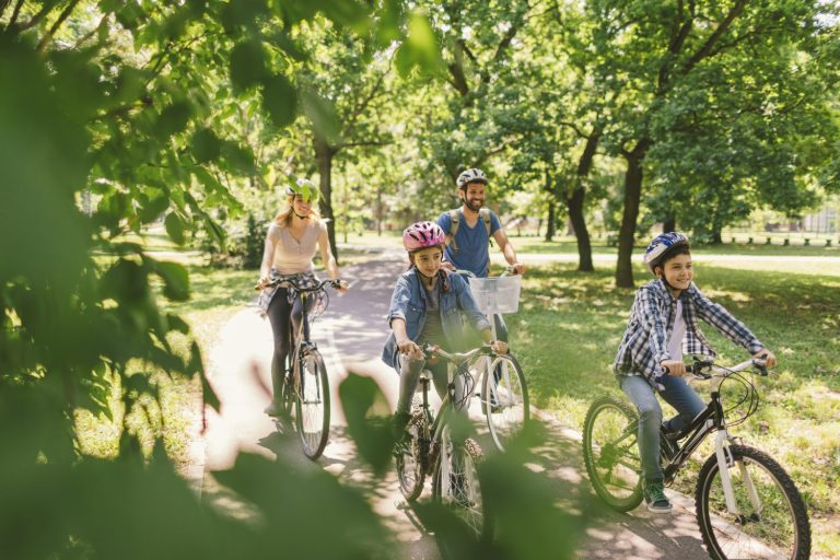 A family of four, wearing helmets, rides bicycles together on a paved path through a green, sunlit park.
