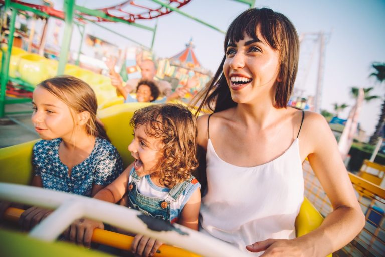 A woman and two children ride a roller coaster at an amusement park, smiling and enjoying the ride on a sunny day.