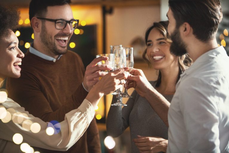 Four people are smiling and clinking glasses together in a celebratory toast at an indoor gathering with warm lighting and blurred string lights in the background.