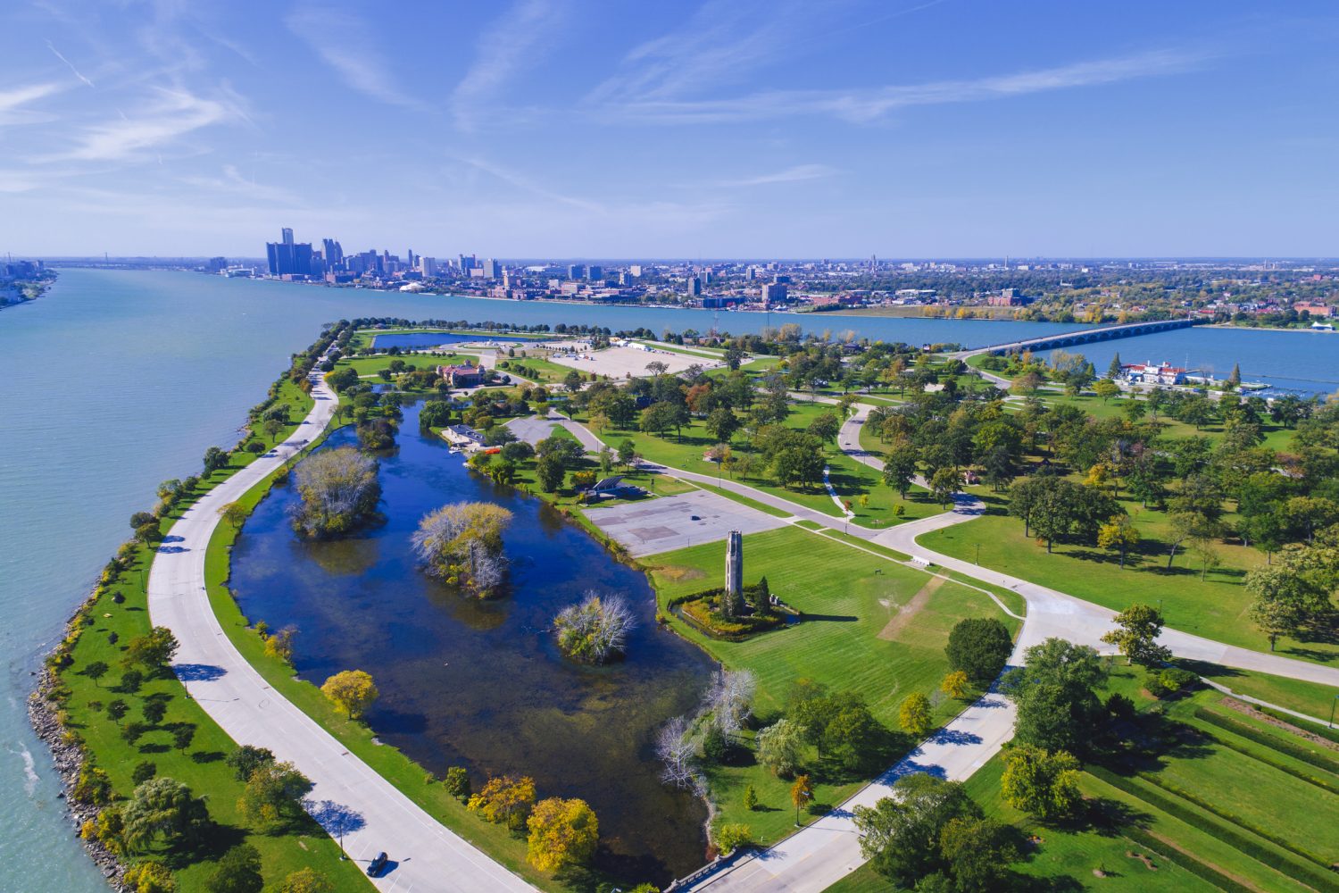 Aerial view of a green park with roads, trees, and flooded areas, bordered by a wide river and a city skyline visible in the background.