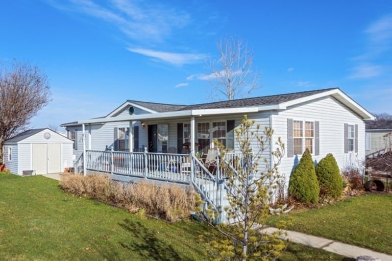 Single-story white manufactured home with a covered front porch, surrounded by grass, shrubs, and a small shed in the backyard under a clear blue sky.