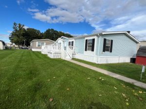 Light blue manufactured home with porch, white trim, and green lawn under a partly cloudy sky.