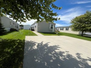 A manufactured home with a large driveway, shaded by trees, on a sunny day in a neighborhood.