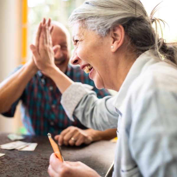 Two older adults sit at a table playing cards, smiling and giving each other a high-five.