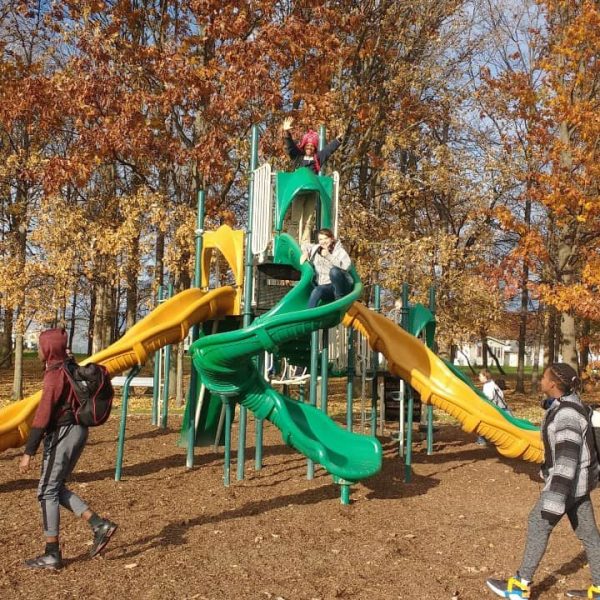 Children play on a green and yellow playground slide structure surrounded by autumn trees, while two people with backpacks walk by.