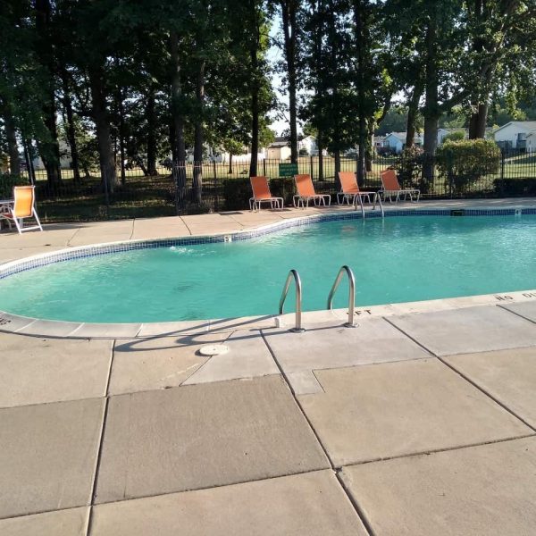 Outdoor swimming pool with metal ladder, surrounded by a concrete deck and orange lounge chairs; trees and houses visible in the background.
