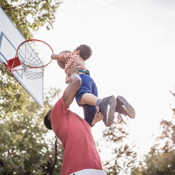 An adult lifts a child toward a basketball hoop outdoors, helping the child reach the rim on a sunny day.